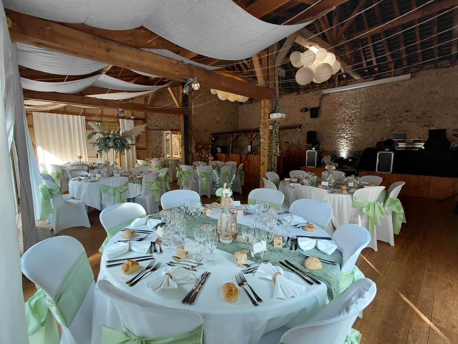 La grande salle dressée pour un mariage : tables rondes nappées de blanc, nœuds vert tendre sur les chaises
